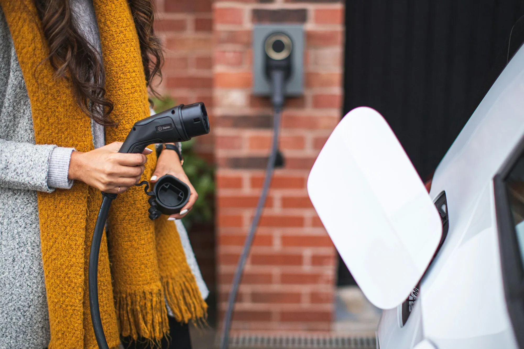 Person in a yellow scarf lifting a charging plug toward a white car